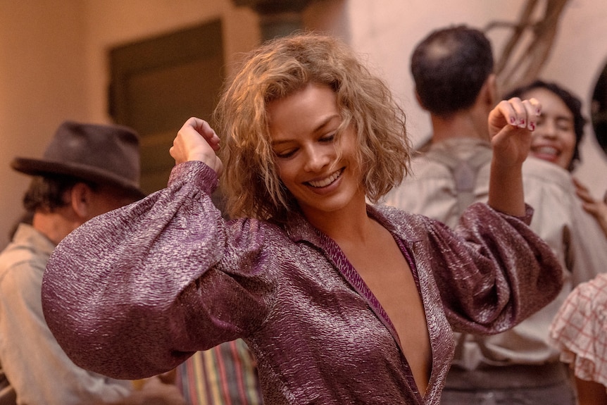 Margot Robbie wears purple dress and smiles while dancing in still from movie Babylon