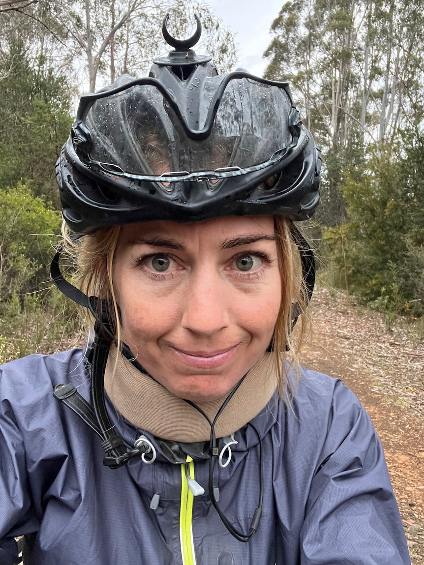 Une femme sourit à la caméra avec une minerve.  C'est un selfie.  Elle porte un casque de vélo noir.