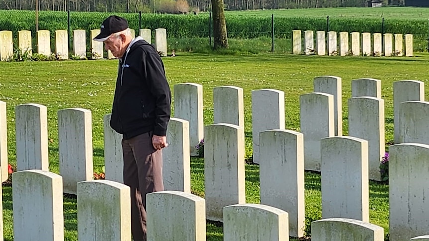 A man stands in a black coat and looks at a tombstone amidst rows of graves