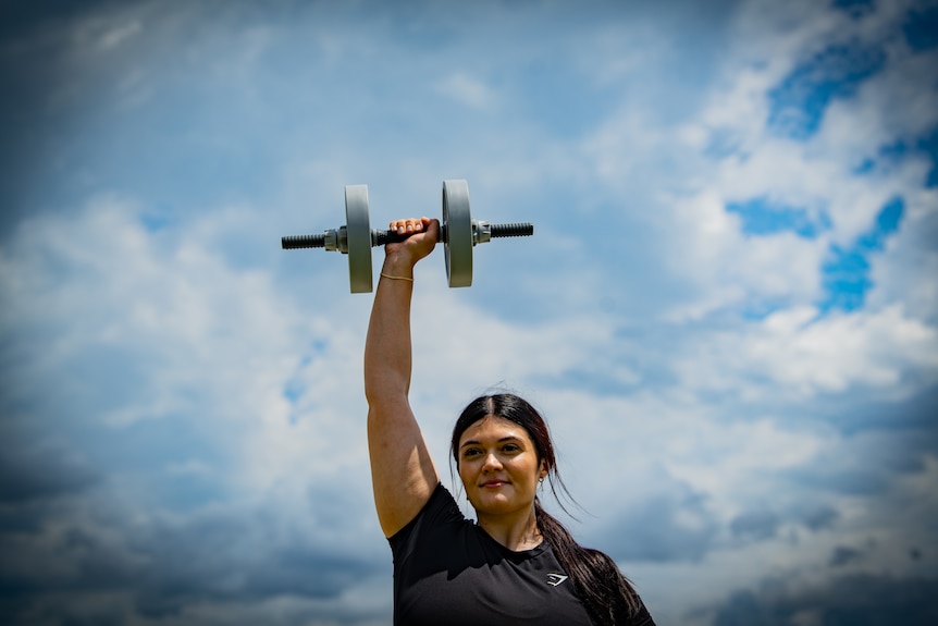 Xanthia holding a single weight above her head, showing larger upper arms, against a cloudy sky.