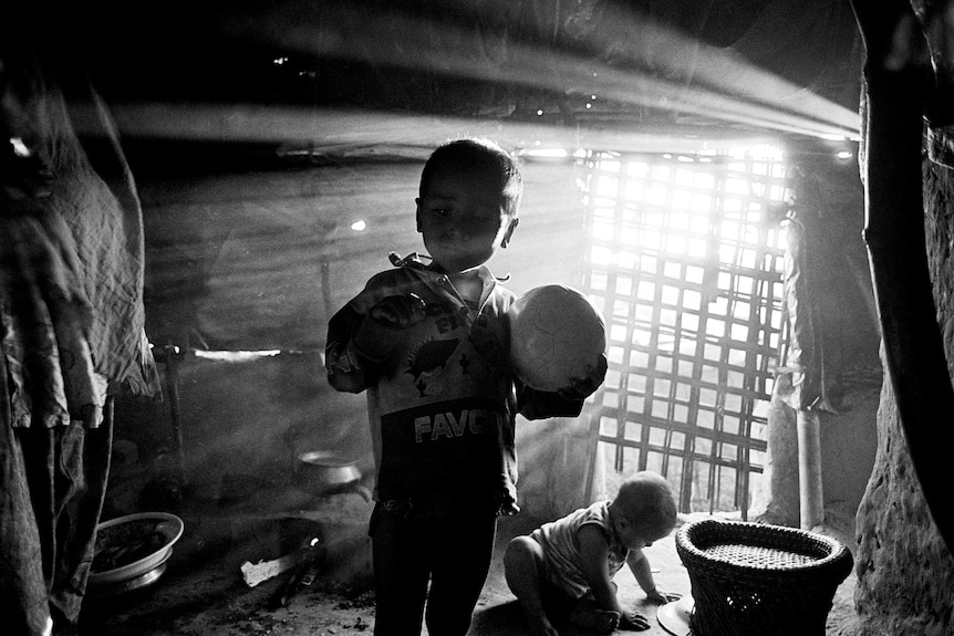 Light streams through the window of a small hut as a Rohingya child holds a soccer ball.