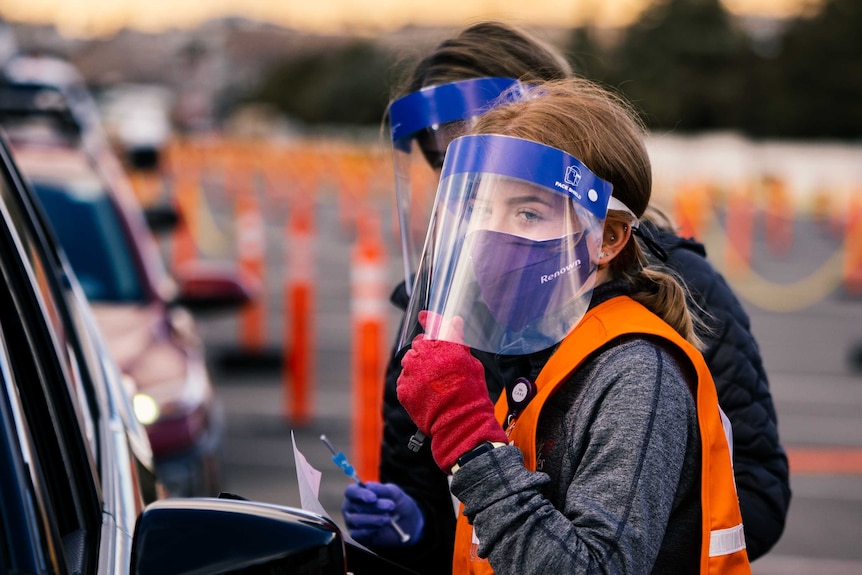Two women in face masks holding needles near a row of cars