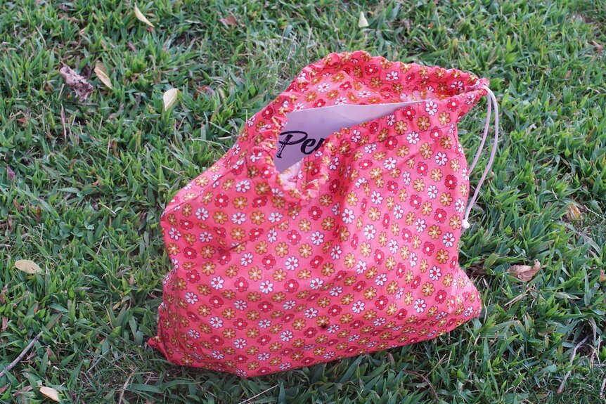 A pink and yellow floral bag is seen resting on grass, with a piece of paper poking out of it.