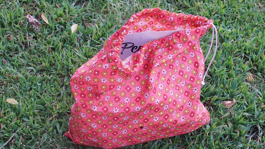 A pink and yellow floral bag is seen resting on grass, with a piece of paper poking out of it.