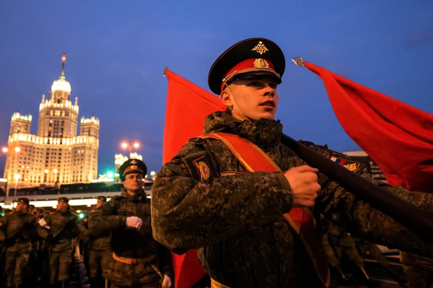 A soldier dressed in military uniform and wearing a cap holds a flag while marching with other men.