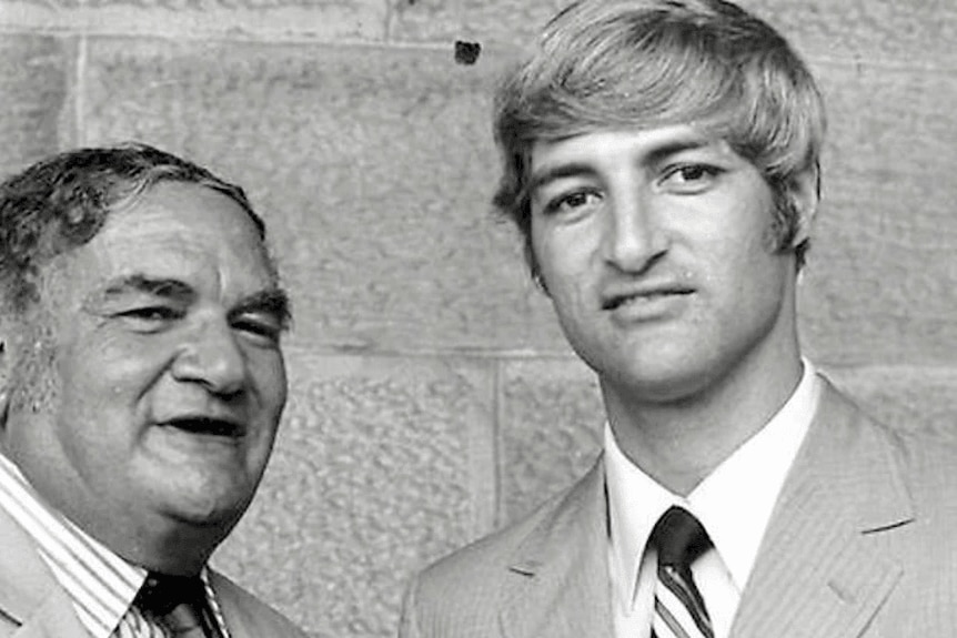 Older man wearing a suit next to his son, a young man also wearing a suit
