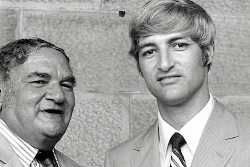 Older man wearing suit next to his son, a young man also wearing a suit