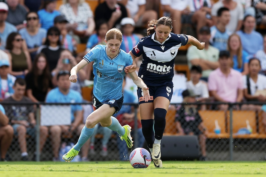 two soccer players, one wearing light blue and the other wearing navy blue, chase the ball during a game with a crowd in the 