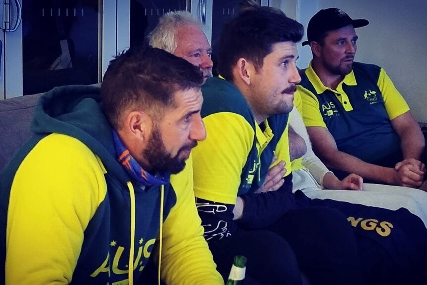 Four men with black hair, wearing green and yellow tracksuits, sit forward on a couch