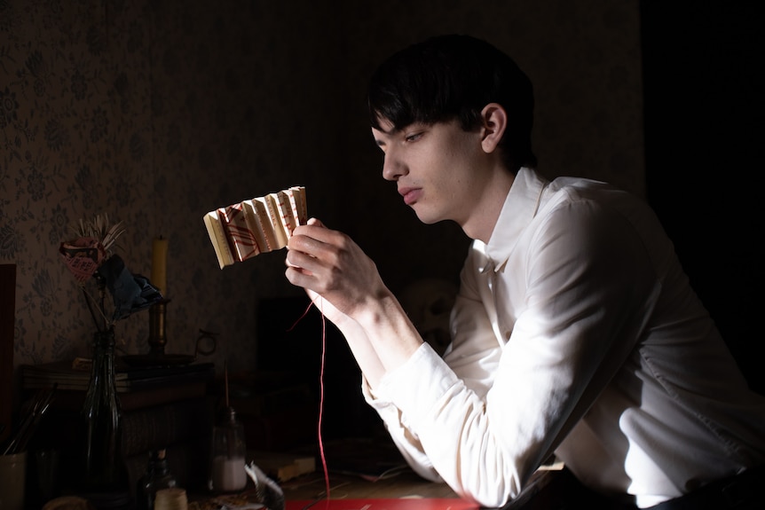 An effete teenage boy in a white collared shirt concentrates as he sits making a paper flower arrangement