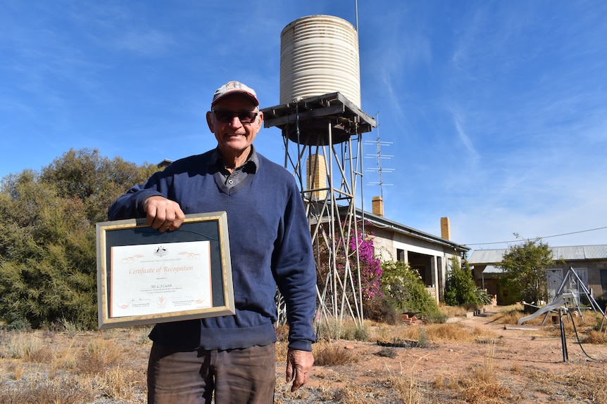 A smiling older man holds a certificate while he stands near an old farmhouse.