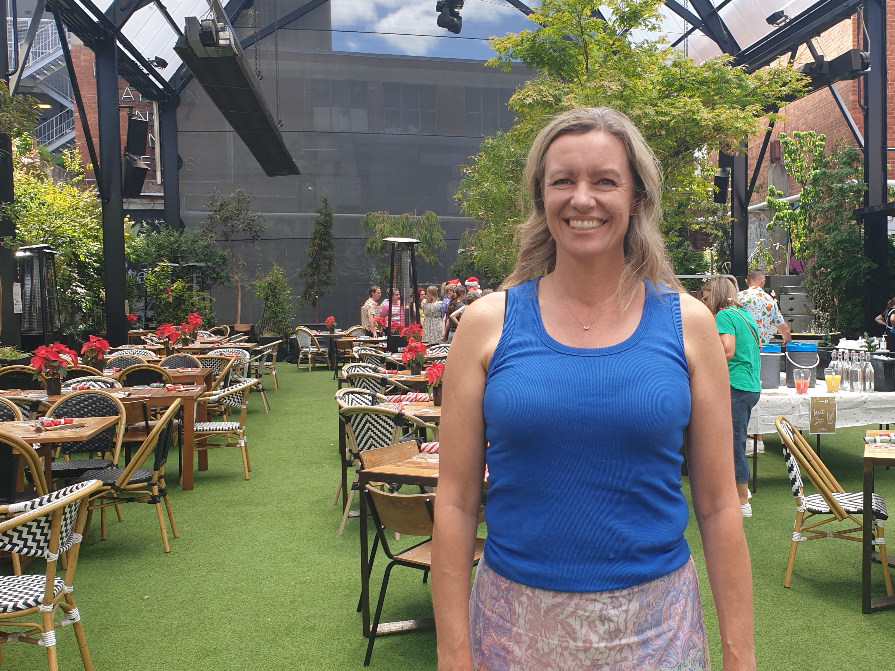 A woman wearing a blue tank standing on a lawn filled with tables and chairs in the background