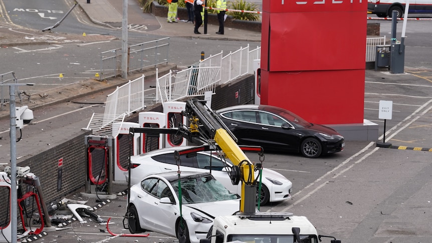 A yellow crane lifts a white Tesla which has a broken windscreen. 