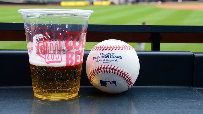 A baseball and cup of beer with a baseball stadium in the background.