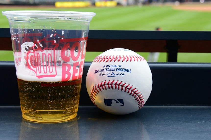 A baseball and cup of beer with a baseball stadium in the background.