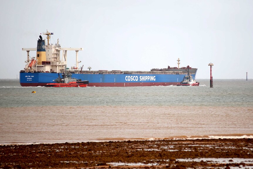A large cargo ship with two pilot boats alongside, under grey skies.