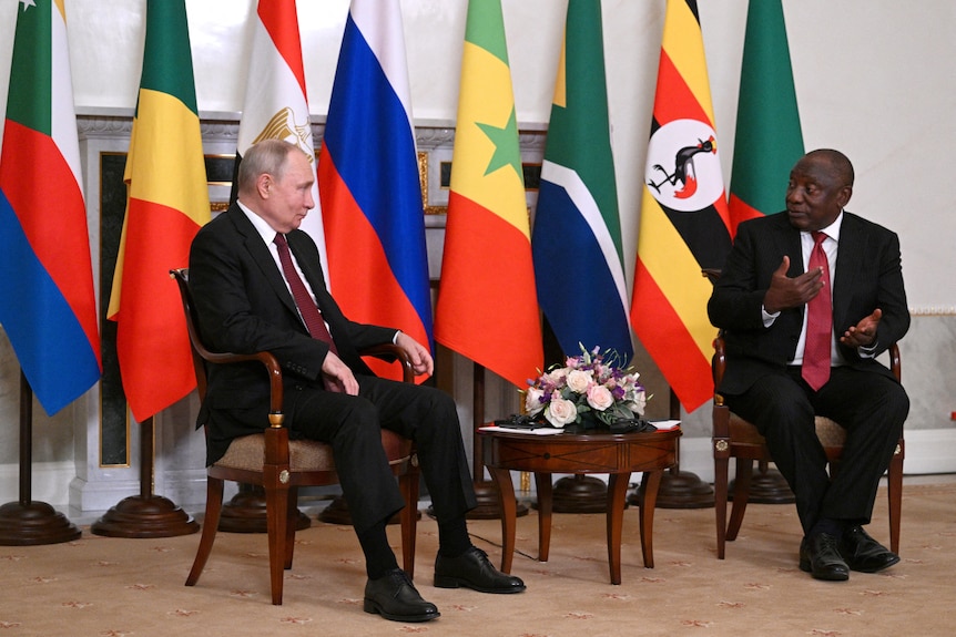 Two men in black suits sit on either side of  small table, in front of a row of flags.