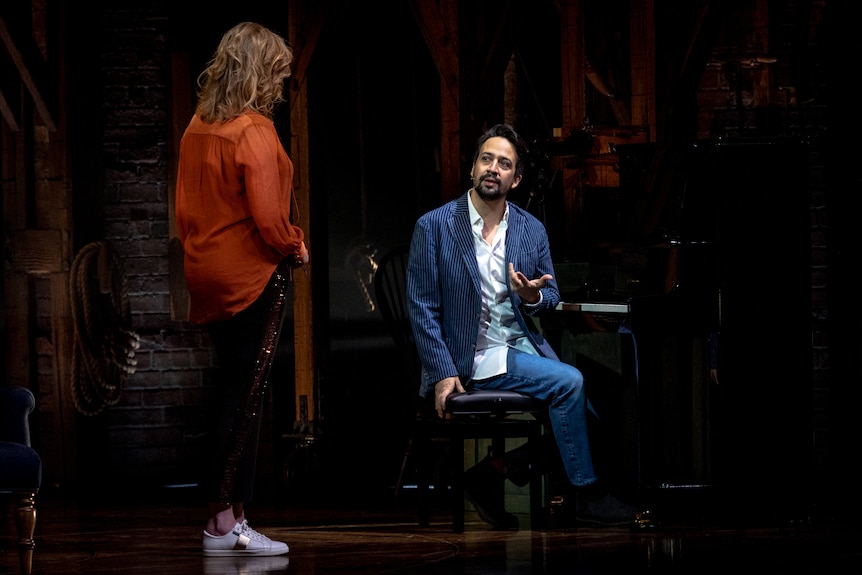 Lin-Manuel Miranda, who's wearing a blue jacket and sitting at a piano, speaks to a woman wearing an orange shirt