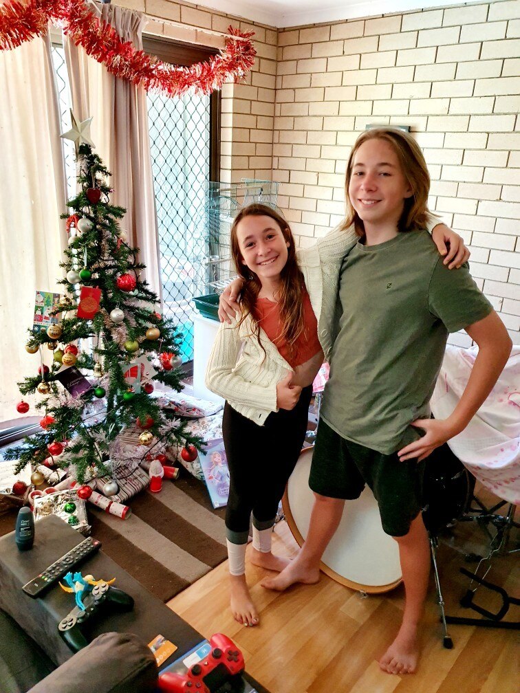 A girl and boy standing inside a home against a brick wall with Christmas tree in the background