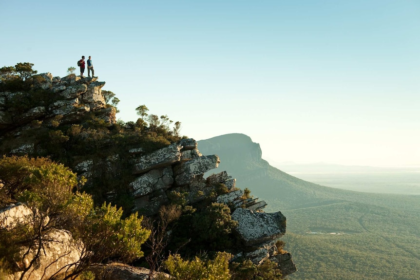 Two people stand on top of a rocky bluff, looking out over a spectacular, mountainous landscape.