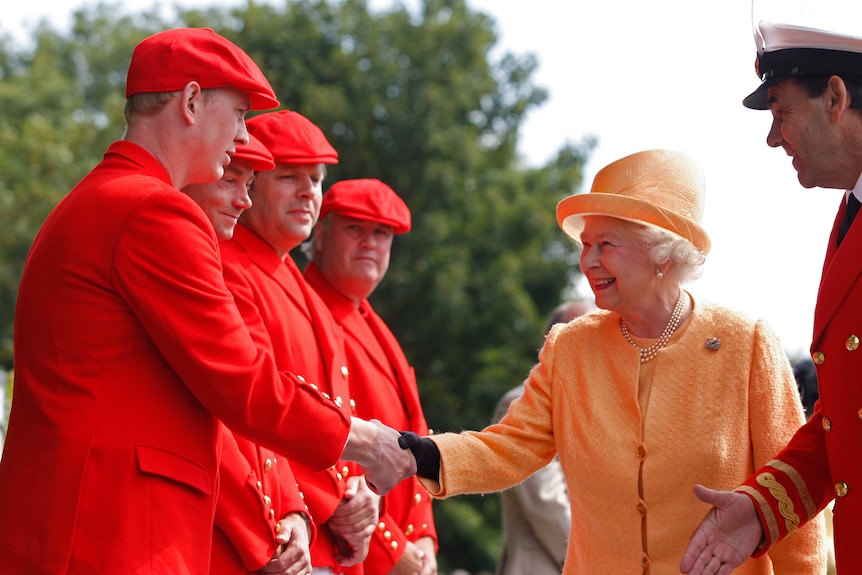 An old lady dressed in orange regal wear shakes hands with men dressed in scarlet rowing clothes