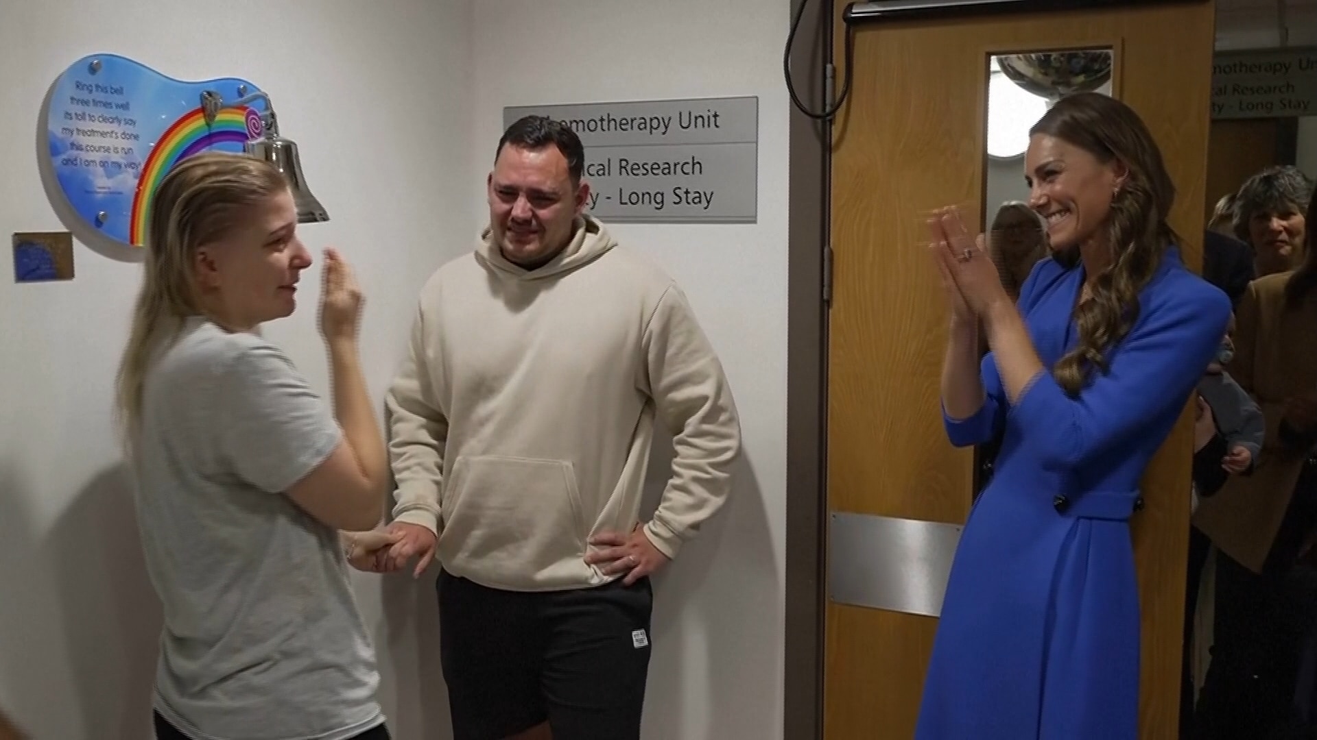 Princess of Wales wearing a blue dress clapping. Standing with a man and woman hugging in a Chemotherapy unit. 