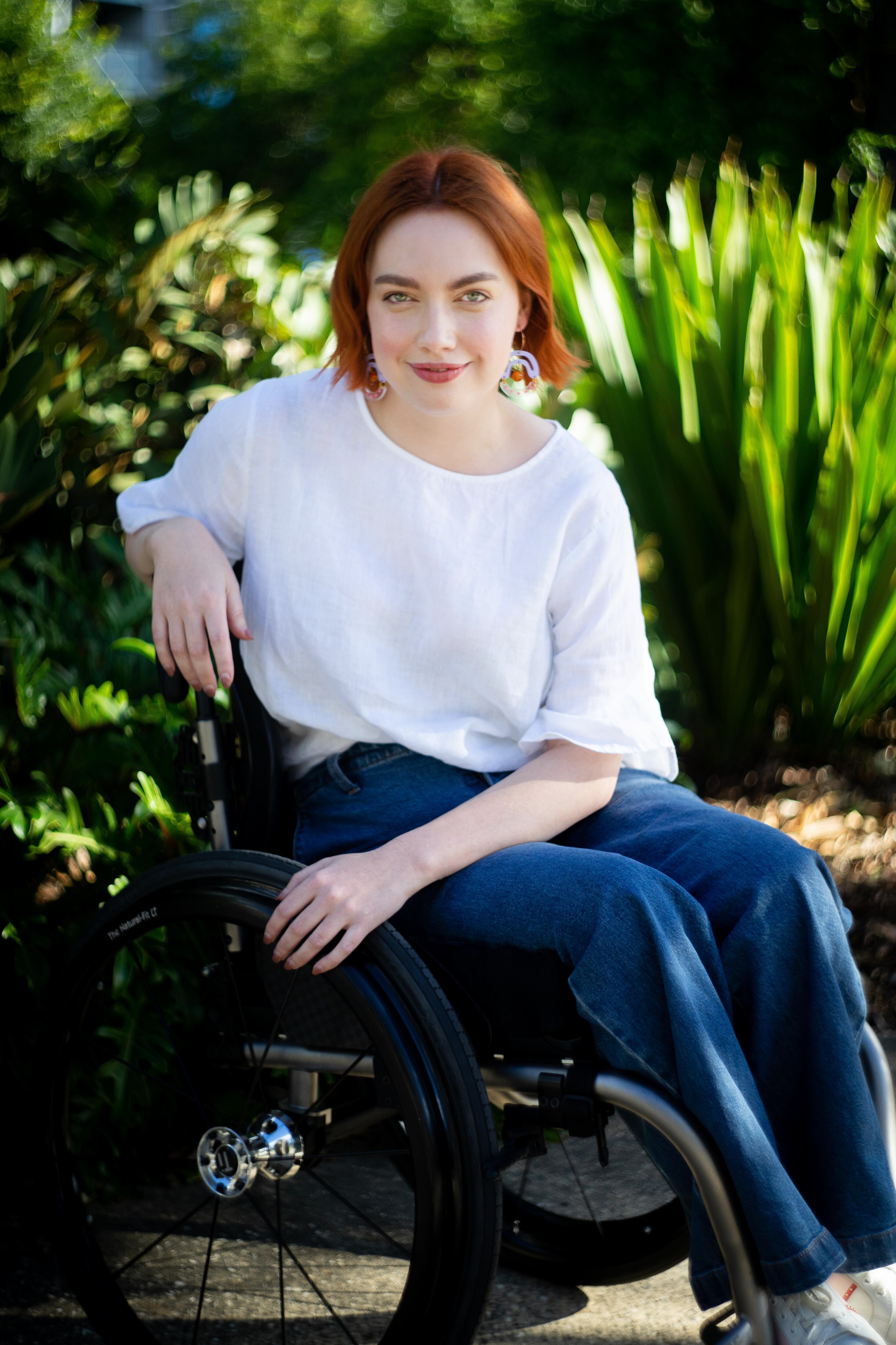 Elly is sitting in her wheelchair, resting her right arm on the back of the chair. She is smiling and has short red hair.