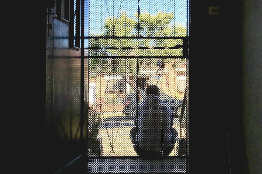 Photo through the front door looking out of John sitting on his front step with back to camera