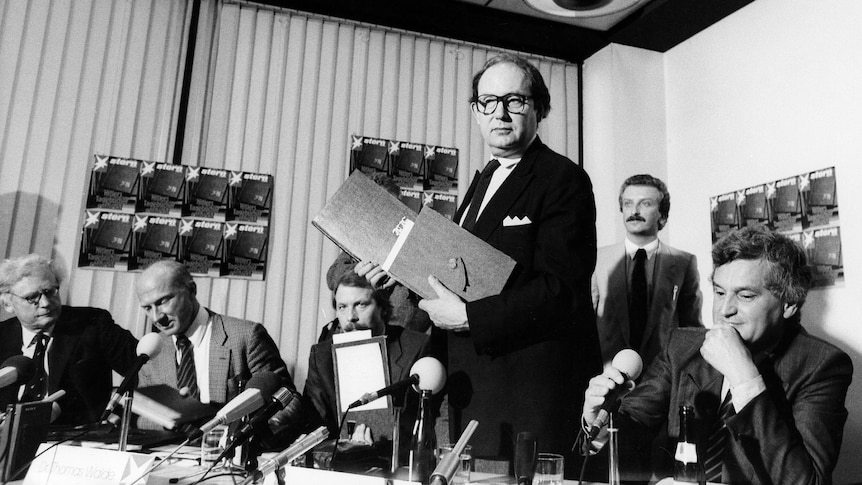 Black and white photo of Gerd Heidemann standing, holding two large books, before group of men seated near microphones.