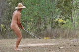 A man wearing only a hat and thongs plays golf in the bush