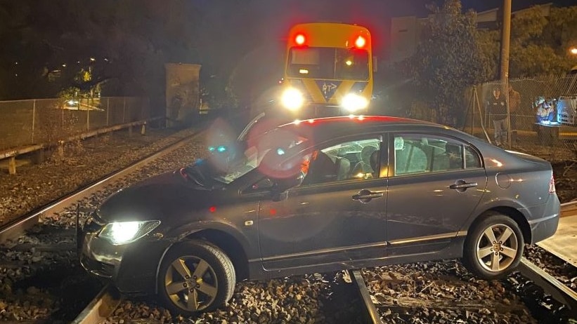 A grey sedan is parked on train tracks at night. A train with headlights on is stopped a few metres behind it.