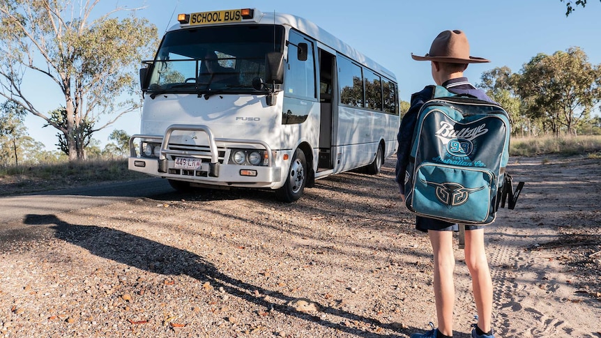 A student with a big backpack waits for a bus on a dirt road
