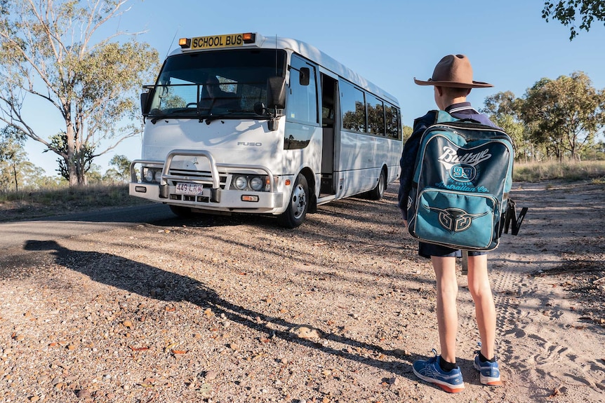 A student with a big backpack waits for a bus on a dirt road