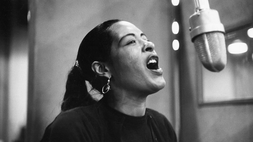A black and white photo of Billie Holiday singing into a microphone. Her hair is a in a ponytail and her mouth is wide open.