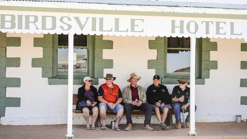 Four men and a woman, all wearing hats, sit drinking on a patio under an awning with the words Birdsville Hotel.