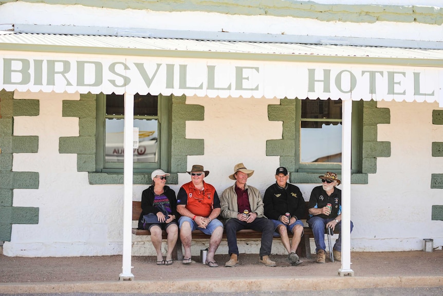 Four men and a woman, all wearing hats, sit drinking on a patio under an awning with the words Birdsville Hotel.