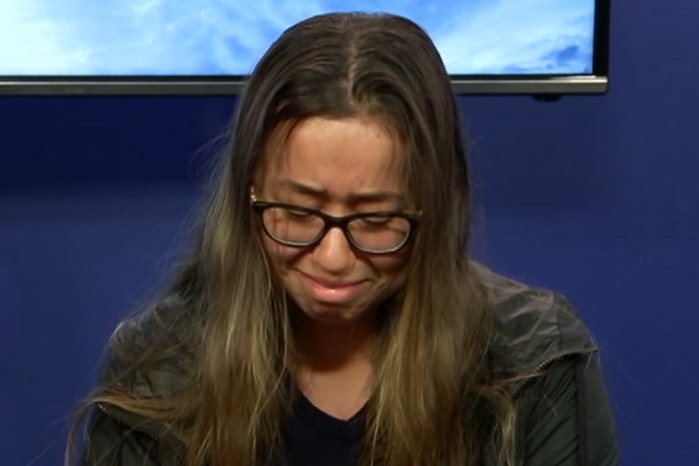 A woman sits in a chair wearing glasses looking down and crying.