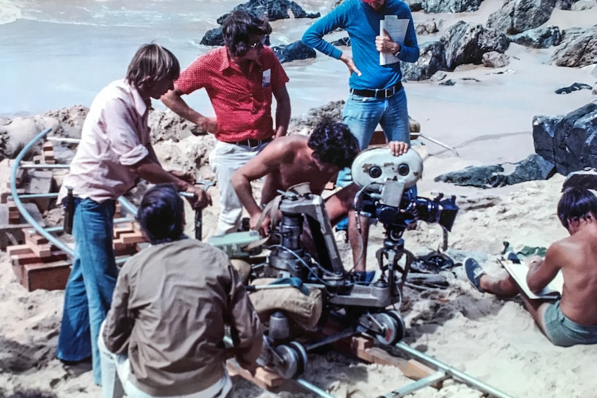 A man with shirts and a bare chest stands behind a camera filming at a beach, as other people look on, in the 1970s.