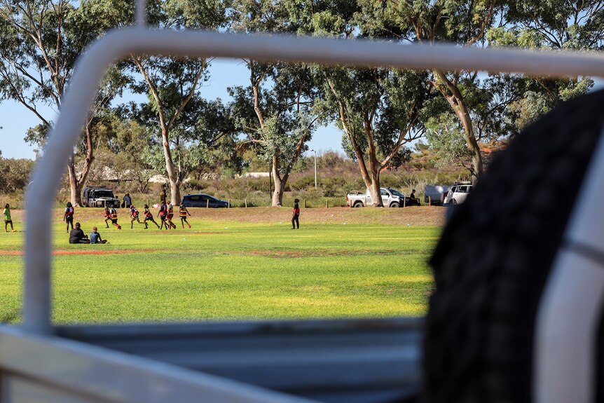 A football ground with young Aboriginal women running viewed through the frame of a white ute truck