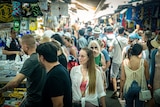 A young woman in a white shirt and pink crop top walks through a packed outdoor marketplace 