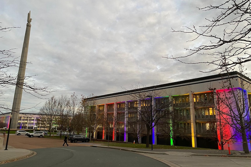 A public building with tall columns lit up with various colours of the rainbow, a similarly lit building visible behind.
