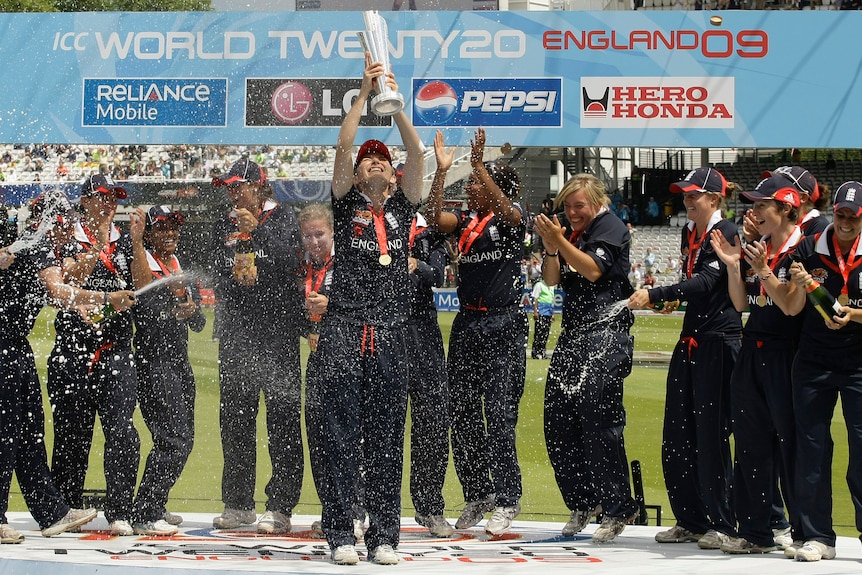 A cricketer holds a trophy aloft in the centre of the image. Her teammates stand behind he cheering and spraying champagne.