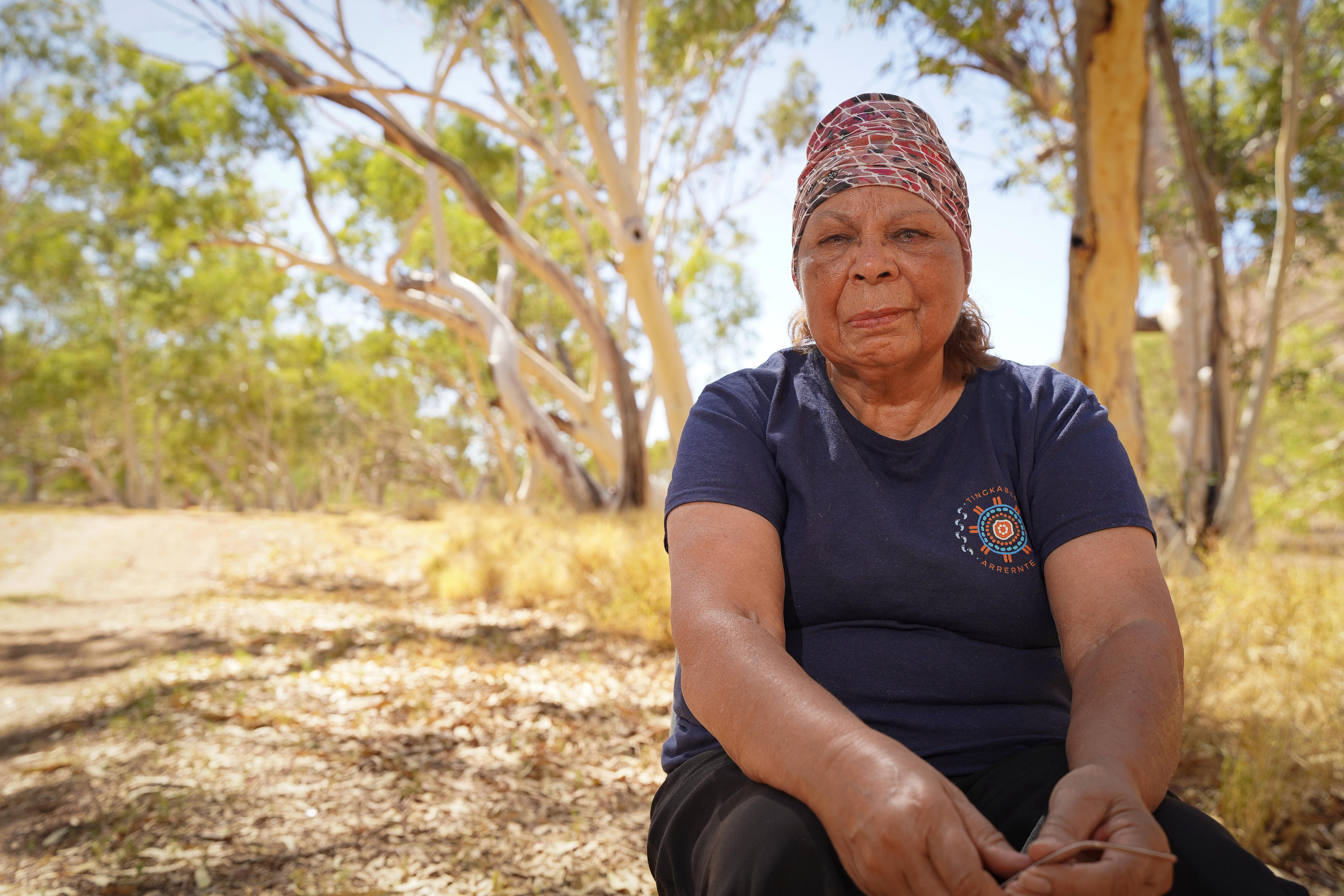 An Aboriginal woman, wearing a patterned head scarf, navy blue top sitting in a bushland area.