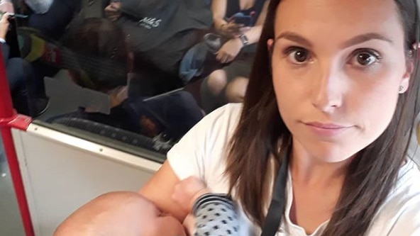 A mother looks at the camera angry while she stands and breastfeeds her baby on a busy train.