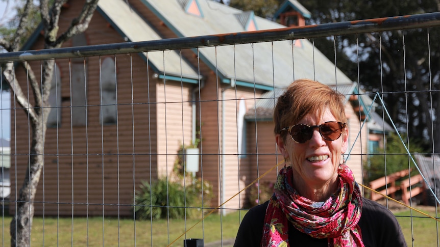 An older woman with red hair , sunglasses and a bright pink scarf stands in front of a fenced-off pink church. 