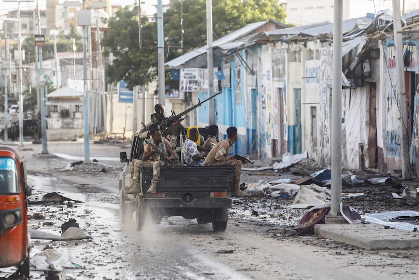 Somali security forces with rifles sit in the back of a truck as it drives along street littered with debris.