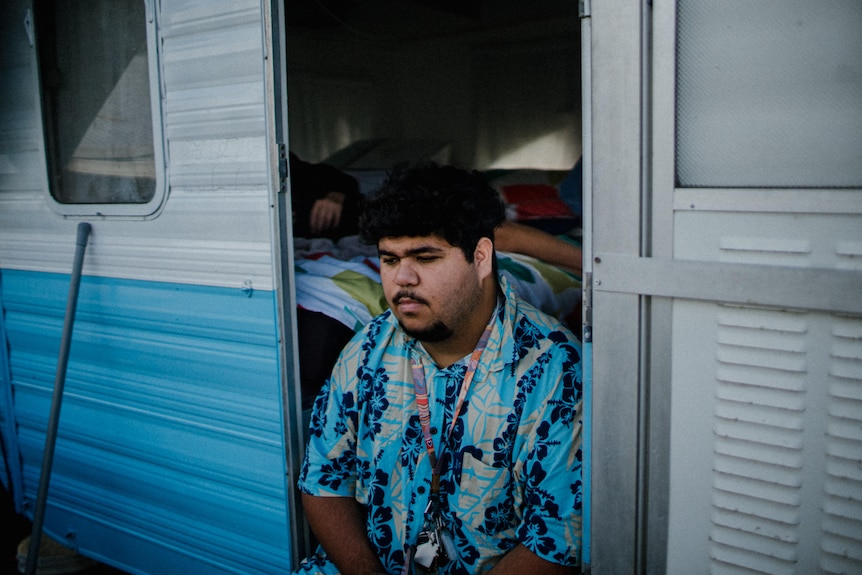 Jesiah sits on the doorstep of a caravan, looking downwards.