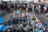At least 100 people are seen in the background standing around blue and white candles on the ground with flowers.