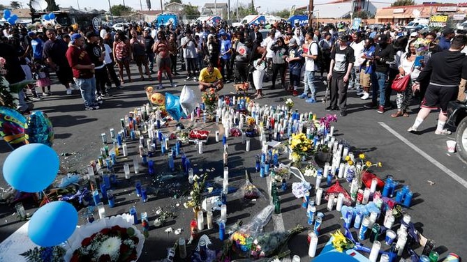 At least 100 people are seen in the background standing around blue and white candles on the ground with flowers.
