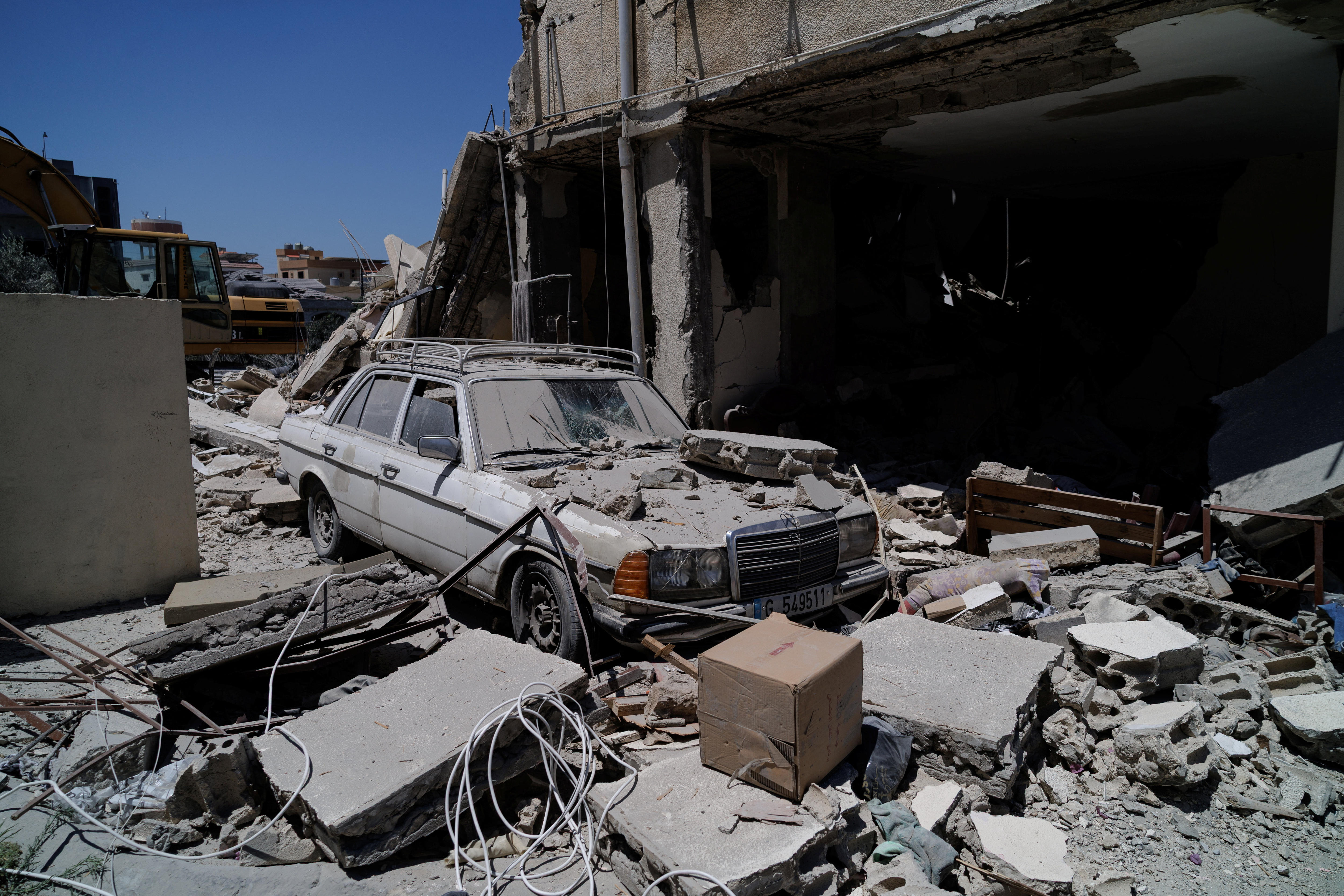 A white car is covered in rubble as it stands next to a badly damaged house.